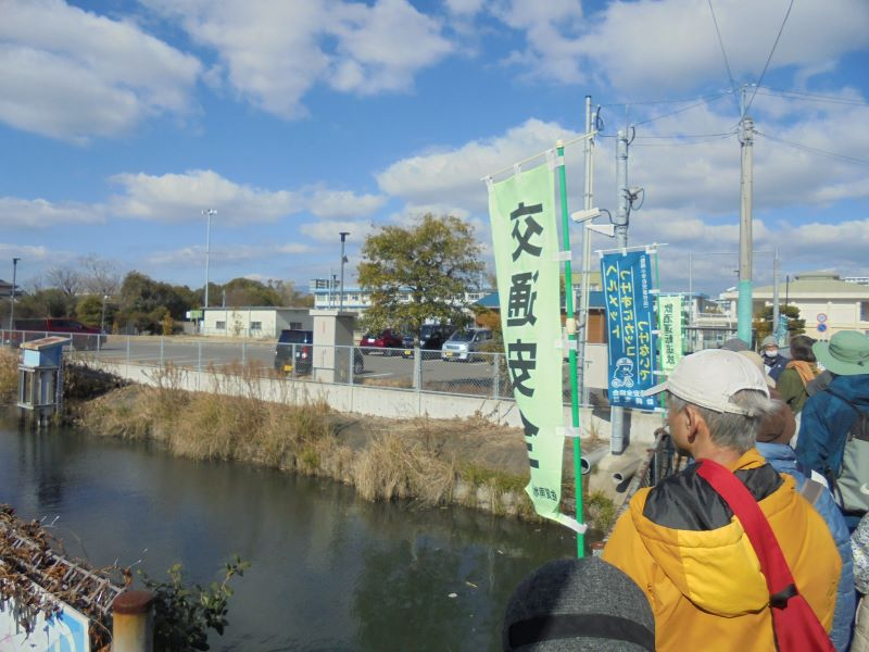 川沿いの堤防で集まる参加者の背後に交通安全の旗と駐車場越しの建物が広がる佐賀市第2水源地跡の写真