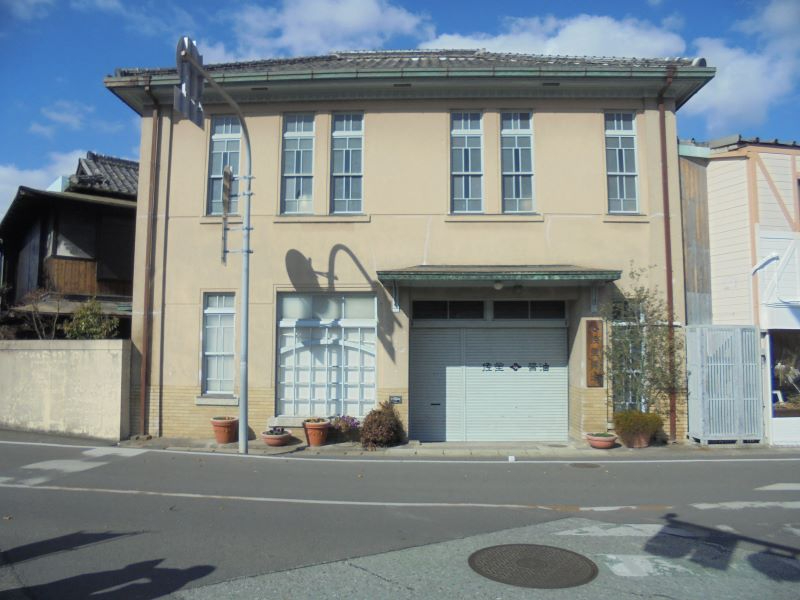 薄茶色の二階建て建物が交差点に面して立ち、並んだ窓と鉢植えが静かな町並みに調和する佐星醤油の店舗兼事務所と居宅を正面から捉えた写真