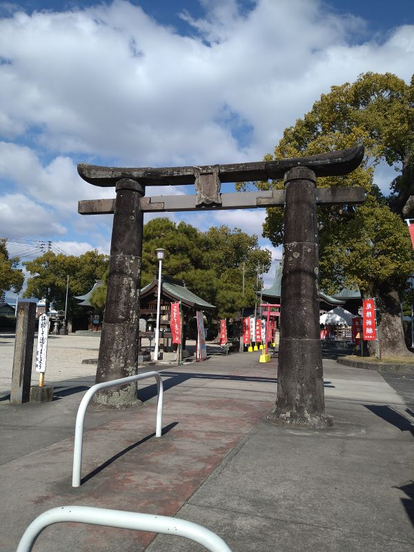 古びた石造りの鳥居の先に拝殿と赤い幟が整然と並び、青空の下で豊かな木々に囲まれた広い神社参道を写した写真