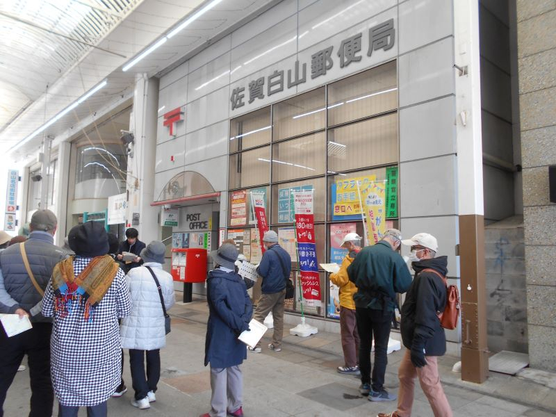 商店街の屋根付き通路沿いにある佐賀白山郵便局前で多数の人々が案内表示を見たり並んだりしている様子を捉えた写真