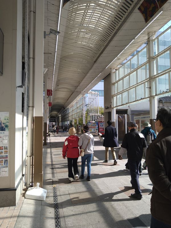 屋根付き白山名商店街の明るい通路を複数の歩行者が日差しを受けながらゆっくり進む様子を捉えた写真