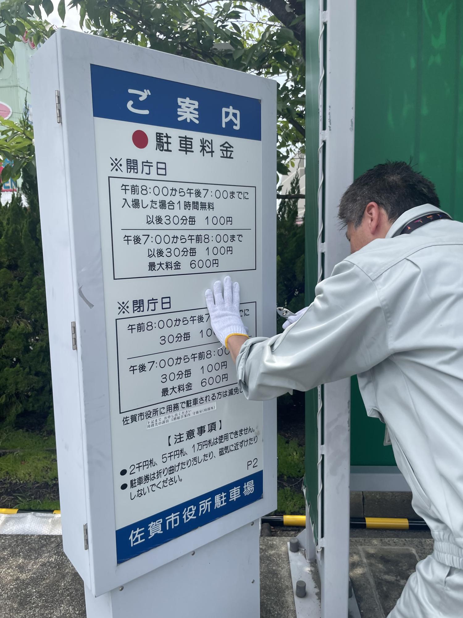 駐車場案内板に料金や開庁日、注意事項が記載され、作業服を着た人物が白手袋で看板を点検している写真