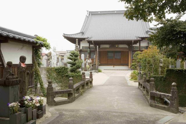 趣ある日本家屋の山門をくぐり抜けた先に、石造りの橋が本堂へと真っ直ぐに伸び、その傍らには静かに見守るような地蔵尊や整えられた庭木が並ぶ、歴史ある寺院の清浄で落ち着いた境内を捉えた写真