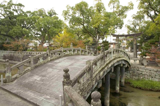 穏やかな水面に映り込む緑豊かな木々に囲まれ、石垣で組まれた境内へと続く風情ある石造りの太鼓橋が架かり、その先には鳥居や石灯籠が静かに佇む、歴史ある神社の情緒豊かな景観を捉えた写真