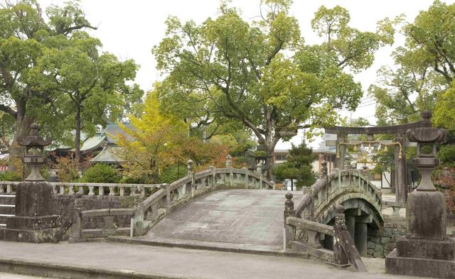 歴史ある神社の境内に向けて、池の上にゆるやかな弧を描く石造りの太鼓橋が架かり、その周囲を深い緑の木々が包み込むように生い茂る、静謐で情緒あふれる風景を捉えた写真