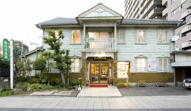 薄いブルーの下見板張りの外壁と瓦屋根が調和した木造2階建ての洋風建築で、中央にバルコニー付きの玄関ポーチを備え、左側には「薮内写真館」と書かれた緑色の看板が立つ、昭和レトロな趣を現代に伝える薮内写真館の外観写真
