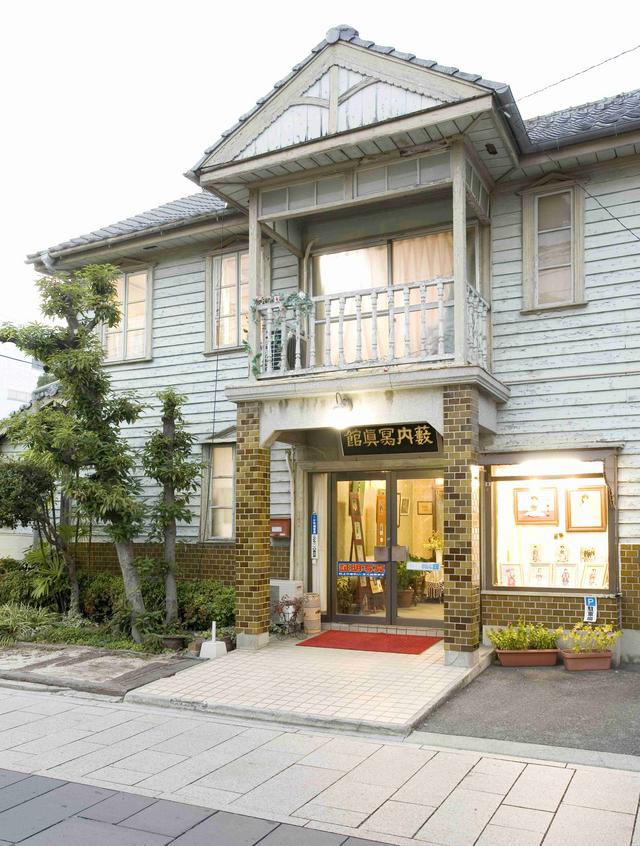 薄いブルーの下見板張りの外壁が特徴的な洋風建築であり、2階部分へと突き出したバルコニーや、タイル装飾が施された玄関ポーチ、そして屋根に掲げられた「薮内寫眞館」の文字がレトロな趣を感じさせる建物の入り口付近を間近に捉えた写真