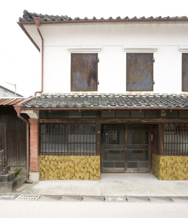 瓦屋根の2階部分に4つの錆びた鉄扉があり、1階には赤レンガの柱と黄色いタイル装飾、格子窓が組み合わさった和洋折衷のデザインが特徴的な、北島邸の正面入口付近を捉えた外観写真