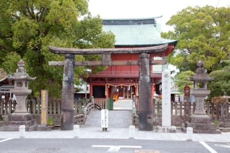 大きな楠の木々に囲まれて、歴史を感じさせる石造りの鳥居が立ち、その奥には朱色の鮮やかな拝殿と太鼓橋が見え、左右には立派な石灯籠と石碑が並ぶ、厳かな神社の風景を捉えた写真