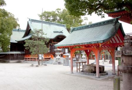 大きな楠の木々に囲まれた境内の中心に、青銅色の屋根と鮮やかな朱色の柱が美しい手水舎が建ち、その奥に重厚な入母屋造りの本殿や拝殿が佇む、歴史ある金立神社の厳かで静謐な雰囲気を伝える風景写真