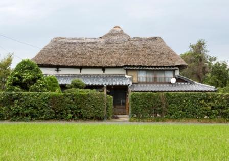 手前に広がる青々とした水田の奥に、厚みのある立派な茅葺き屋根と瓦屋根を組み合わせた伝統的な日本家屋が建ち、周囲を整えられた高い生け垣で囲まれた、日本の原風景を感じさせるのどかな三浦邸の外観写真