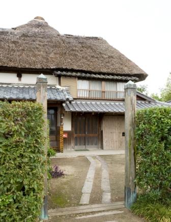 二本の趣ある木柱が立つ門の先に、厚みのある見事な茅葺き屋根と瓦屋根を備えた伝統的な日本家屋が建ち、整えられた生け垣の合間から玄関へと続く石畳の通路が伸びる、歴史情緒溢れる三浦邸の入り口付近を捉えた写真