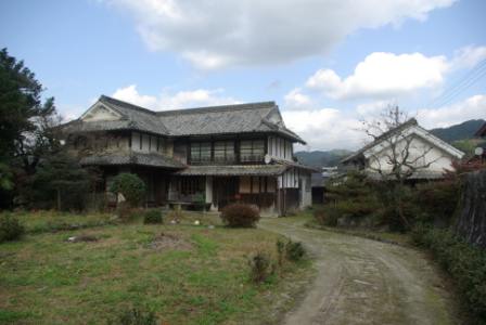前面に草地と曲がった砂利道が広がり、奥に瓦屋根と白壁を持つ日本の伝統的な木造住宅が庭に面して建っている松野邸の写真