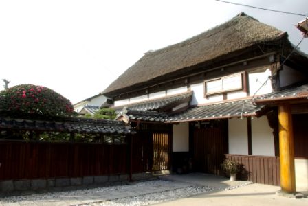 茅葺き屋根を持つ伝統的な日本家屋の門構えで、白壁と木製の格子扉が特徴的で、前面には砂利が敷かれ植木鉢と生垣が配置されている古賀邸の外観写真
