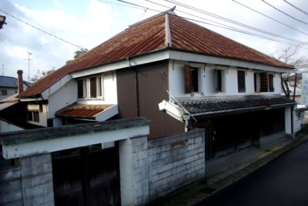 赤茶色に錆びた金属屋根を持つ二階建ての古い日本家屋で、白壁と木製の窓枠が特徴的で、前面には灰色のコンクリート塀と木製門がある千鳥屋総本家原田邸の外観写真