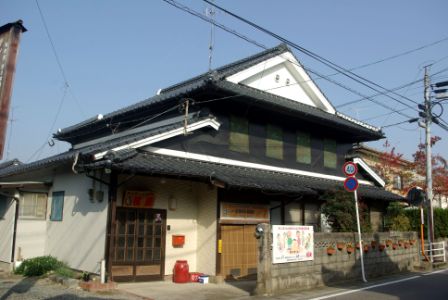 黒い瓦屋根と白壁を持つ日本の伝統的な建物で、正面には木製の引き戸と看板がある横尾邸の外観写真