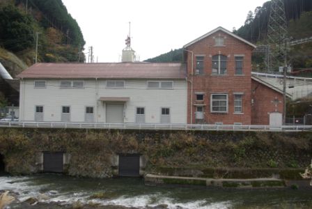 前面に川が流れ、背後に山と送電鉄塔が見える場所に建っている赤いレンガ造りの二階建て部分と白い外壁の平屋部分が連なる九州電力株式会社 川上川第二発電所の写真