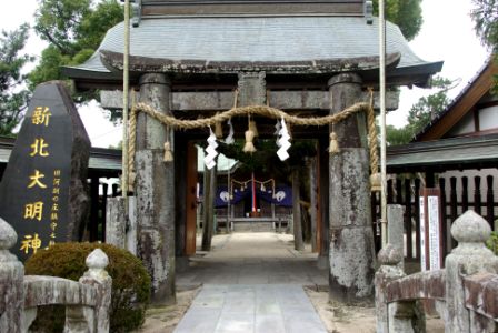 苔むした石柱に注連縄と紙垂が掛けられた鳥居の奥に、紫の幕で飾られた拝殿が見え、左側には「新北大明神」と刻まれた石碑が立つ新北神社の参道の写真