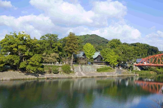 穏やかな川の流れを挟んで、対岸に深い緑の木々に守られるように佇む神社の社殿と、その右手に鮮やかな朱色の鉄橋が架かる、青空と山の稜線が美しい自然豊かな風景を捉えた写真