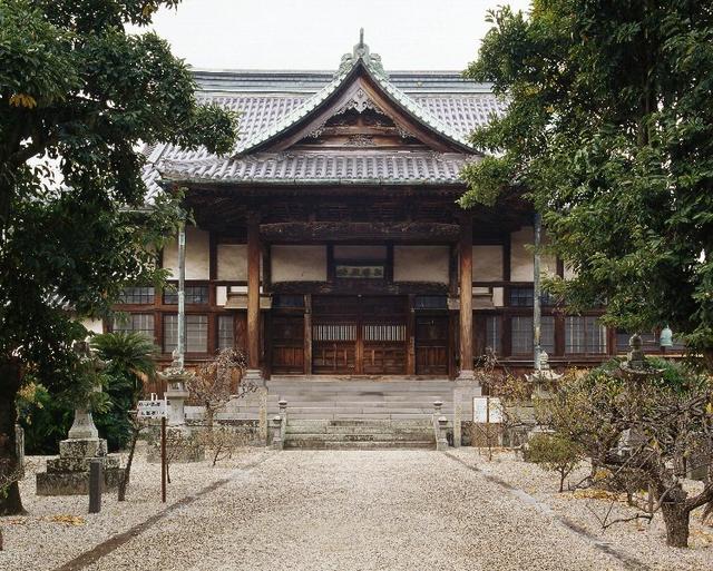 瓦屋根と木造の構造が特徴的な日本の伝統的寺院で、正面に彫刻装飾のある玄関と広い石段があり、両側に木々と石灯籠が並ぶ参道が続く高伝寺の写真