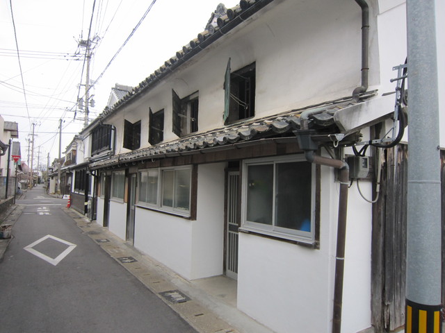 細い路地の脇に建つ白壁の平屋が奥へと続き、2階部分には小さな黒い鉄扉の窓がいくつも並び、1階には新しく改修されたと思われるアルミサッシの窓や引き戸が設置された、南里邸の写真