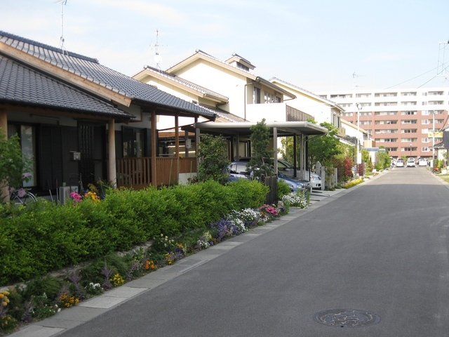 整然と並ぶ戸建て住宅が両側に建ち、前庭には色とりどりの花や緑の植え込みが続き、奥には中層マンションが見える静かな住宅街の舗装道路の写真