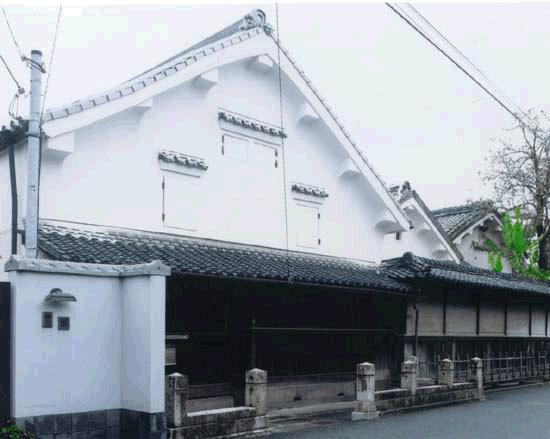 白壁と瓦屋根が特徴的な日本の伝統的な蔵造りの建物で、正面には格子窓と塀が続き、道路沿いに石柱が並ぶ竹下邸の外観写真写真
