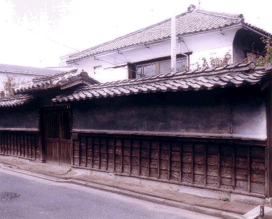 瓦屋根を持つ日本の伝統的な建物で、前面には木製の長い塀と門があ竹下邸の外観写真