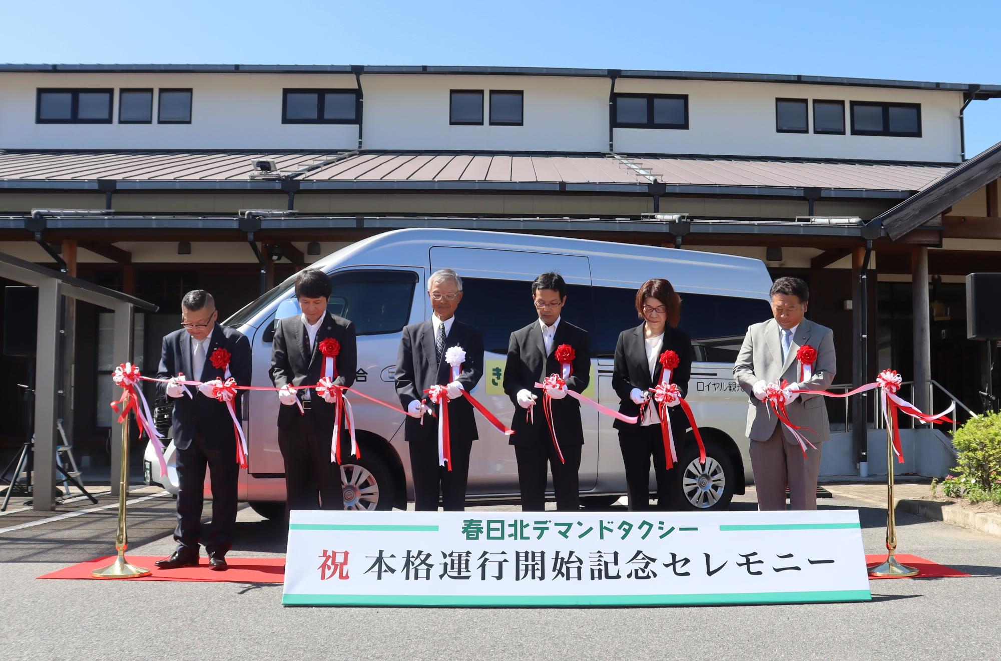 白いワゴン車の前で複数の参加者が赤白のリボンを持ち並んで本格運行開始を祝う式典の様子を写した写真