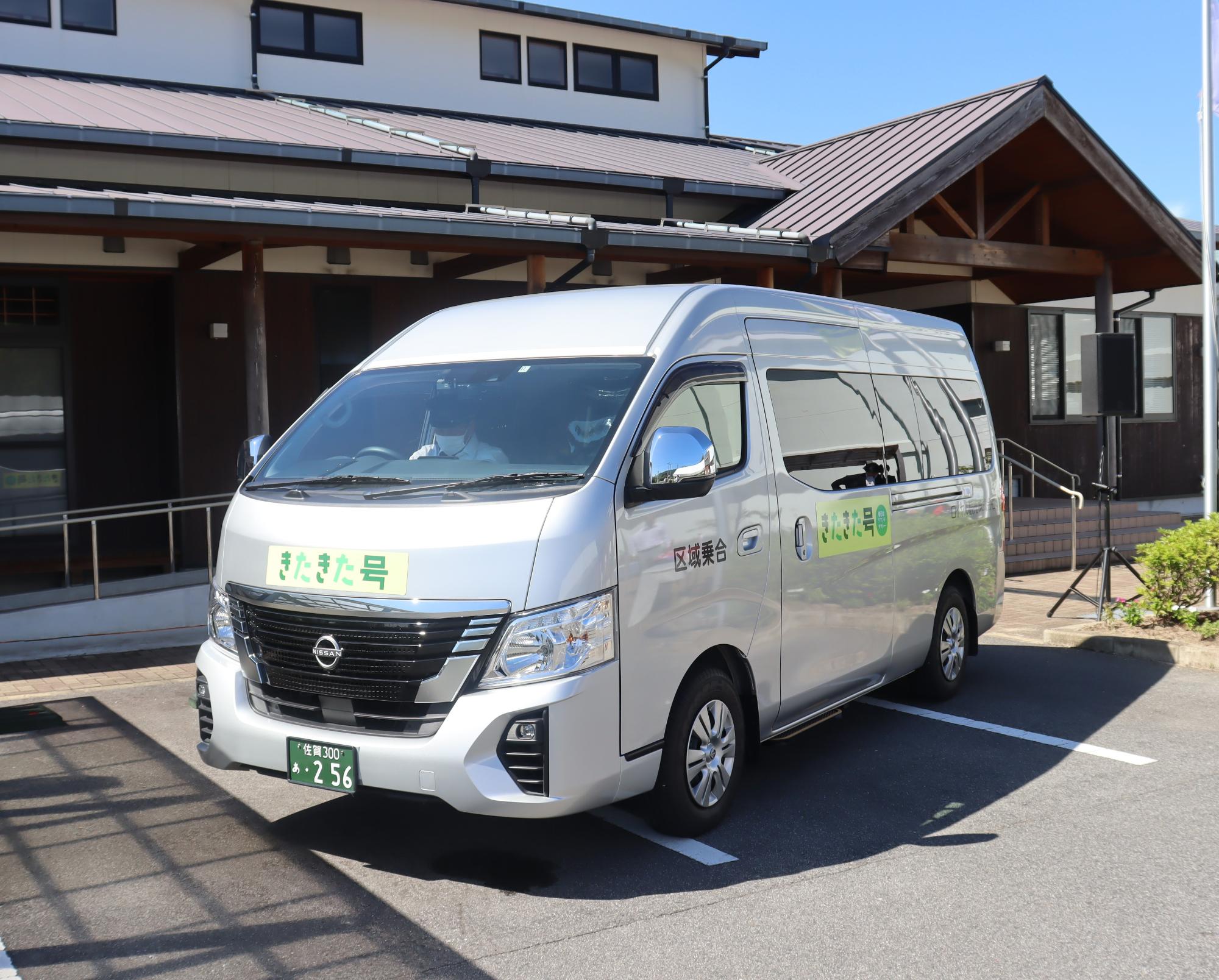 建物前の駐車場に銀色のワゴン車が停まり、側面に緑のきたきた号の案内表示が貼られている様子を写した写真