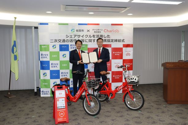 赤いシェア自転車が並ぶ前で二人が協定書を掲げ、連携締結式の会場背景が整然と設置された様子を示す写真