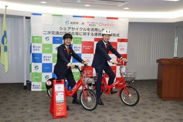 赤いシェア自転車にまたがった二人が連携締結式の会場前で試乗の様子を示している写真