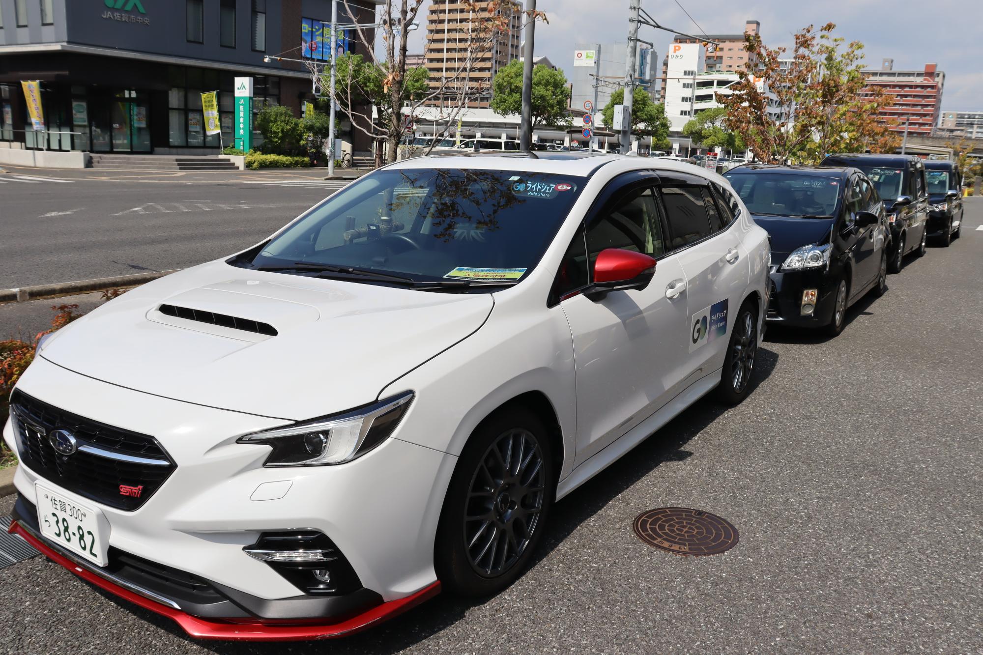 白いライドシェア車両が市街地の道路沿いに黒い車列とともに整然と停車している写真