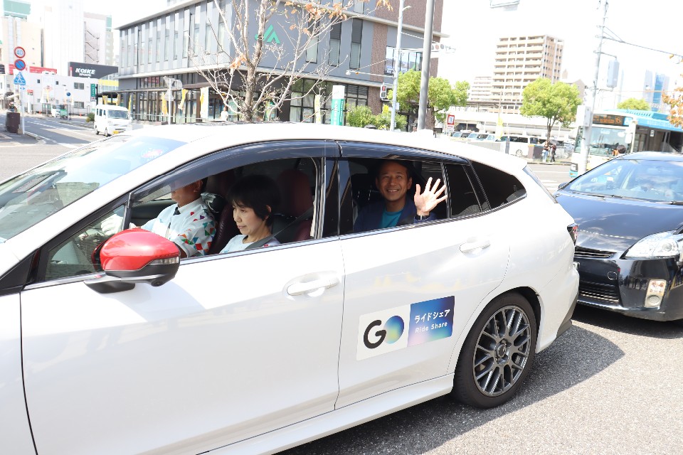 白いライドシェア車両に乗車した後部座席の窓から市長が手を振りながら市街地の道路を走行する様子を捉えた写真