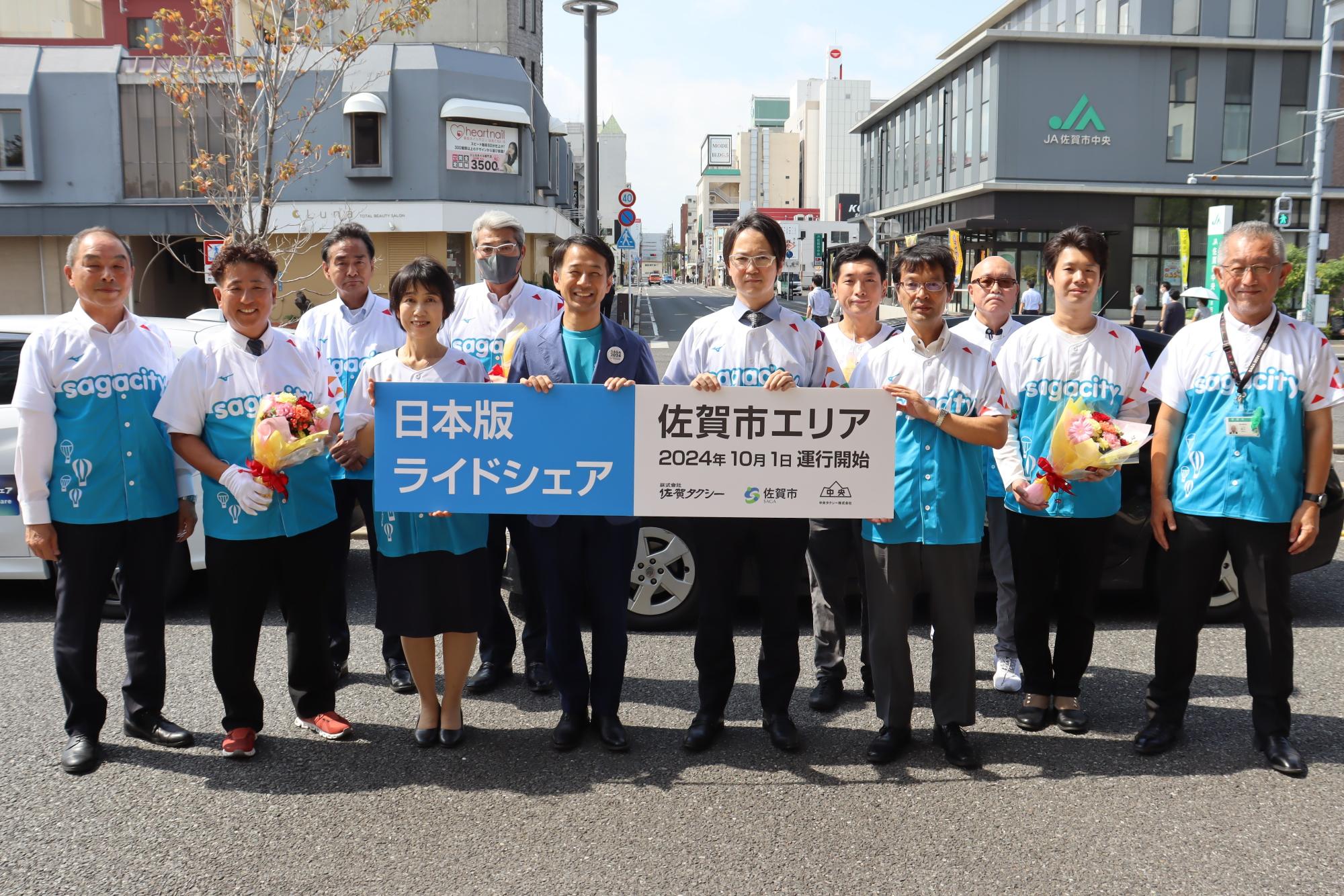 日本版ライドシェア開始を告知する横断幕を持った参加者たちが市街地の車道上に整列して記念撮影する写真