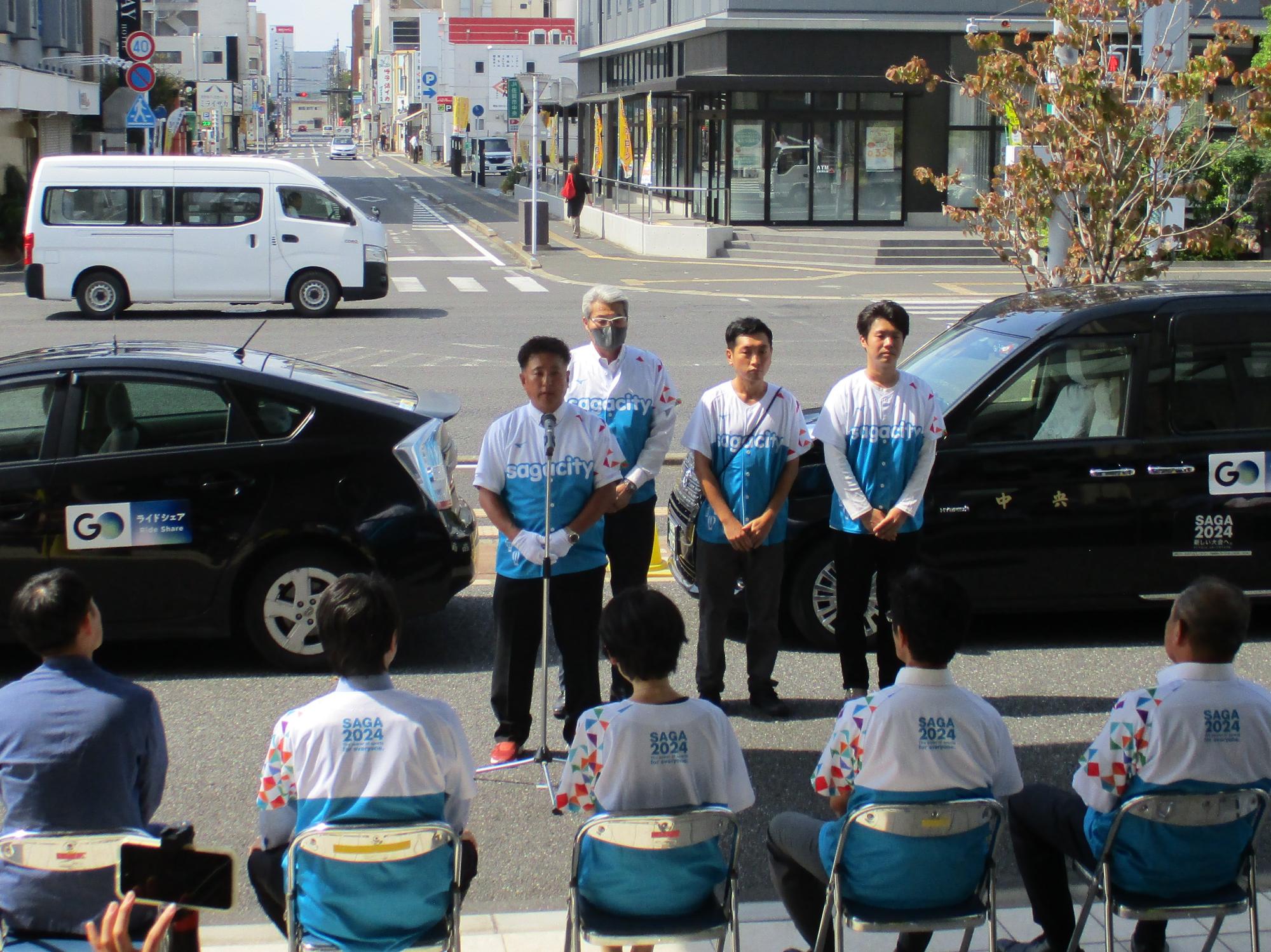 街中の交差点近くでお揃いのユニフォーム姿の参加者が並び、左端に立つ男性が挨拶し、来場者が着席して聞く様子を黒い車両とともに捉えた写真