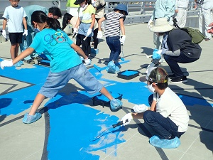 有明海沿岸道路のイベントにて、靴カバーを履いた子どもたちがローラーや筆を使い、コンクリートの路面に引かれた下書きに合わせて鮮やかな青色のペンキを塗り広げている写真