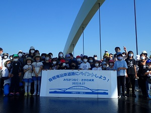 「有明海沿岸道路にペイントしよう！」と書かれた横断幕を囲み、多くの子どもたちや保護者が笑顔で並んで記念撮影をしている写真