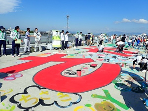 有明海沿岸道路にて、子どもたちが路面に描かれた「さが」などの巨大な赤い文字や、色とりどりの花や雲などの可愛らしいイラストの下書きに合わせて、鮮やかなペンキを使い分けながら彩っている写真