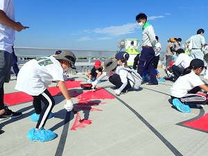 有明海沿岸道路にて、靴カバーを履いた子どもたちが腰を屈めながら、路面に描かれた大きな文字や図形の下書きに合わせて鮮やかな赤色のペンキをローラーで丁寧に塗り広げている写真