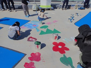 有明海沿岸道路にて、子どもたちが路面に描かれた花や山などの可愛らしい下書きに合わせて、赤や緑、青のペンキを使い分けながら思い思いに色を塗っている写真