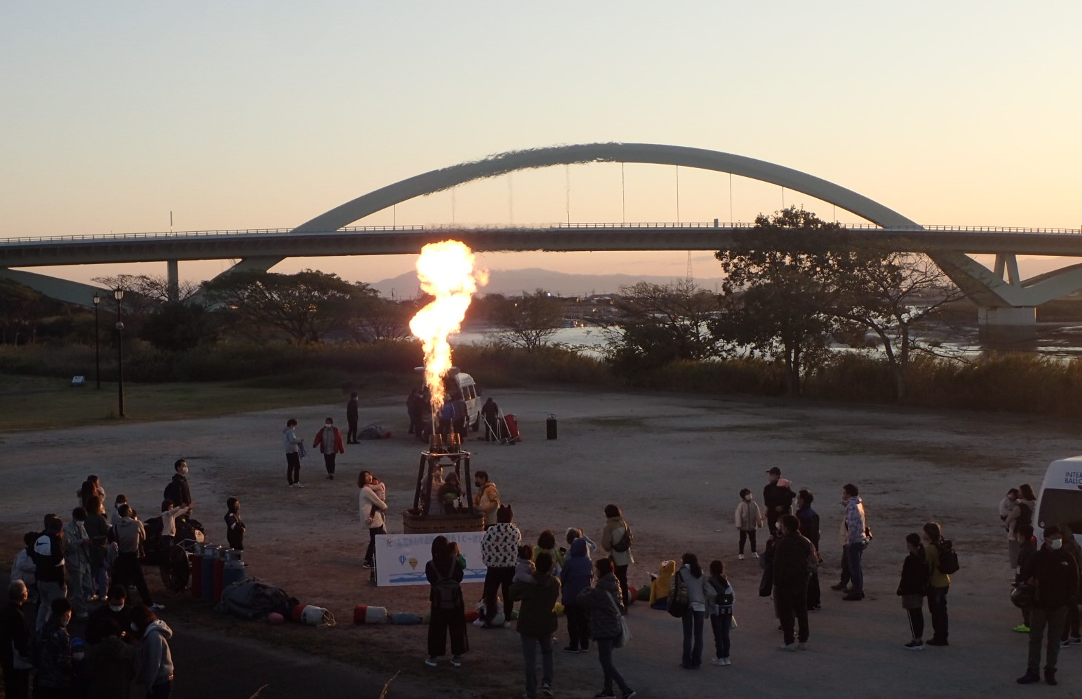 夕暮れの河川敷で大勢の人々が集まり、熱気球のバーナー炎が上がる様子を背後の大きなアーチ橋越しに捉えた写真
