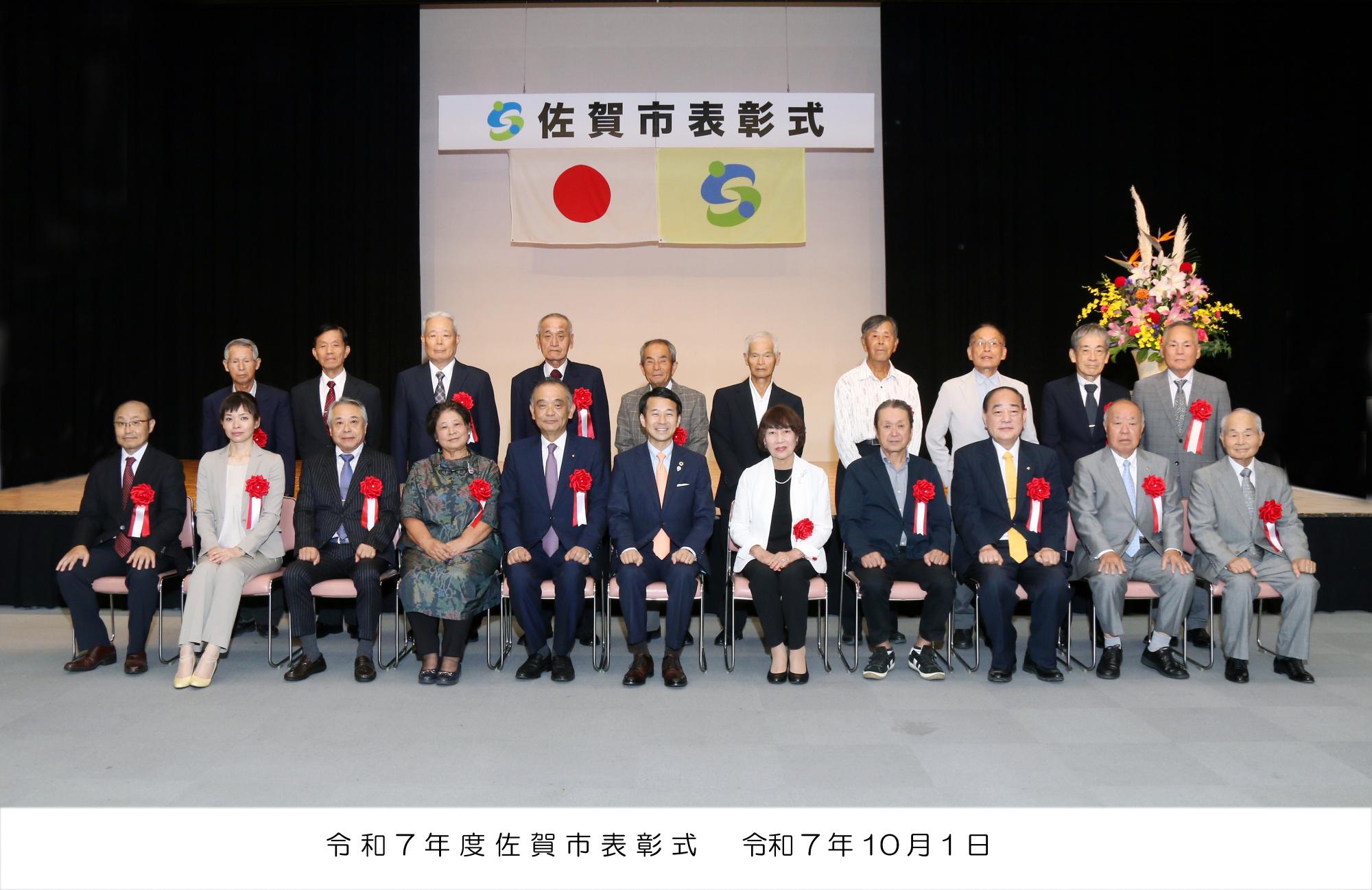 「佐賀市表彰式」と書かれた看板幕と国旗や市旗を背景に、胸に赤いリボンを付けた市長と受賞者の方々が2列に並んで記念撮影を行っている式典の様子の写真