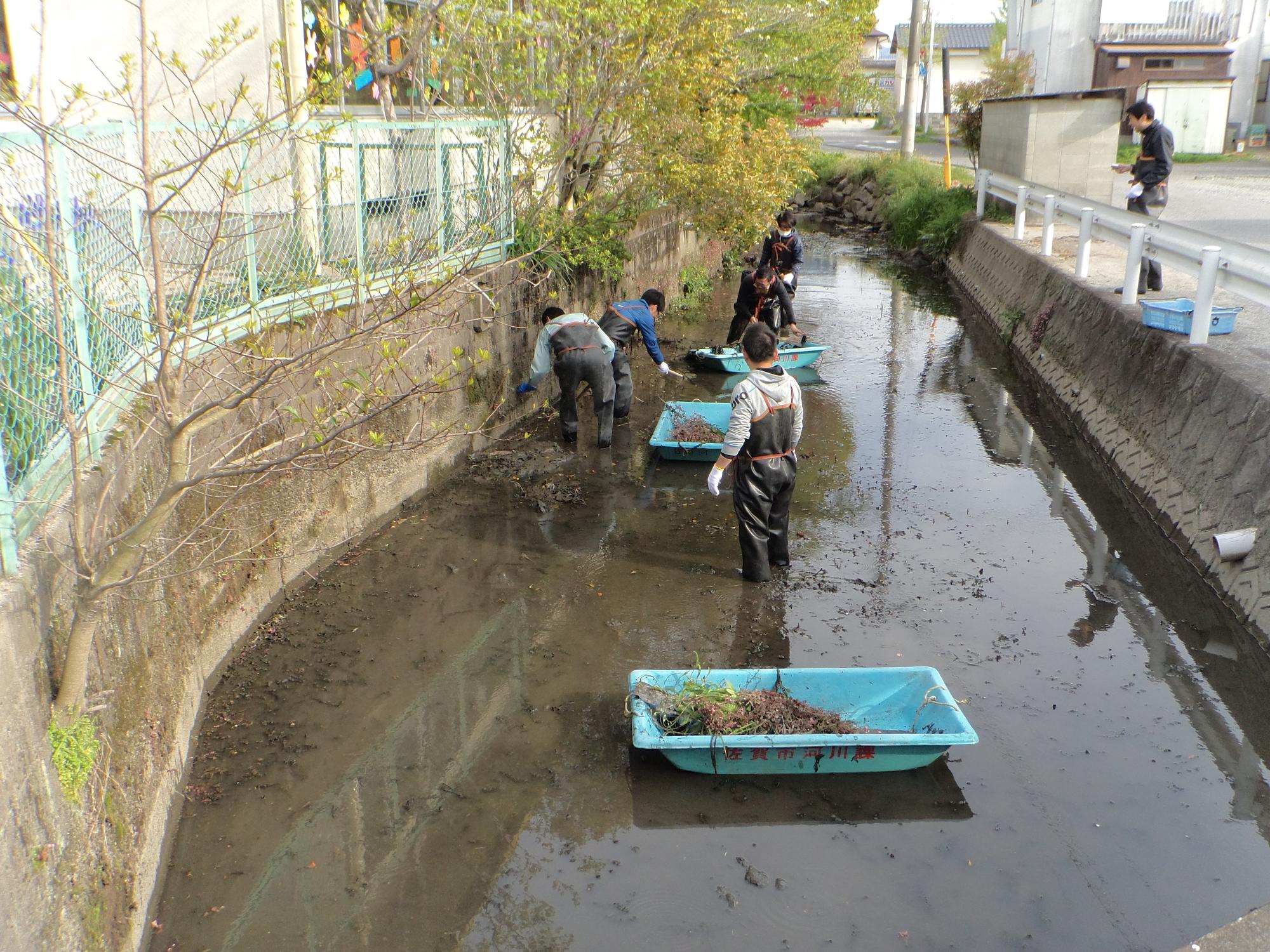 浅い用水路に入った参加者たちが泥や草をすくい取り、手前の舟形容器に集めて清掃作業を進める写真