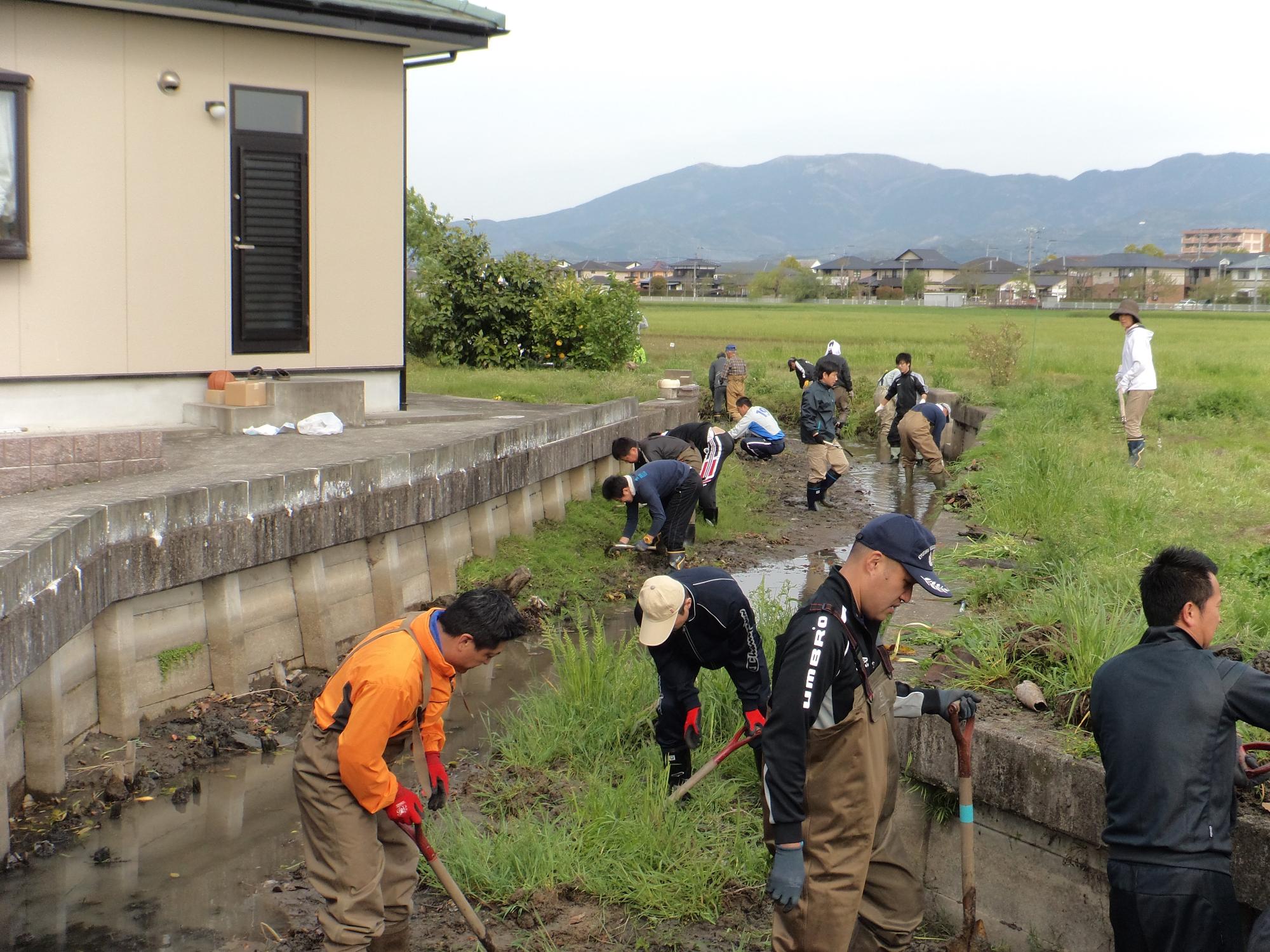農村の水路沿いで作業着姿の参加者たちが泥や草を取り除きながら協力して清掃を行っている写真