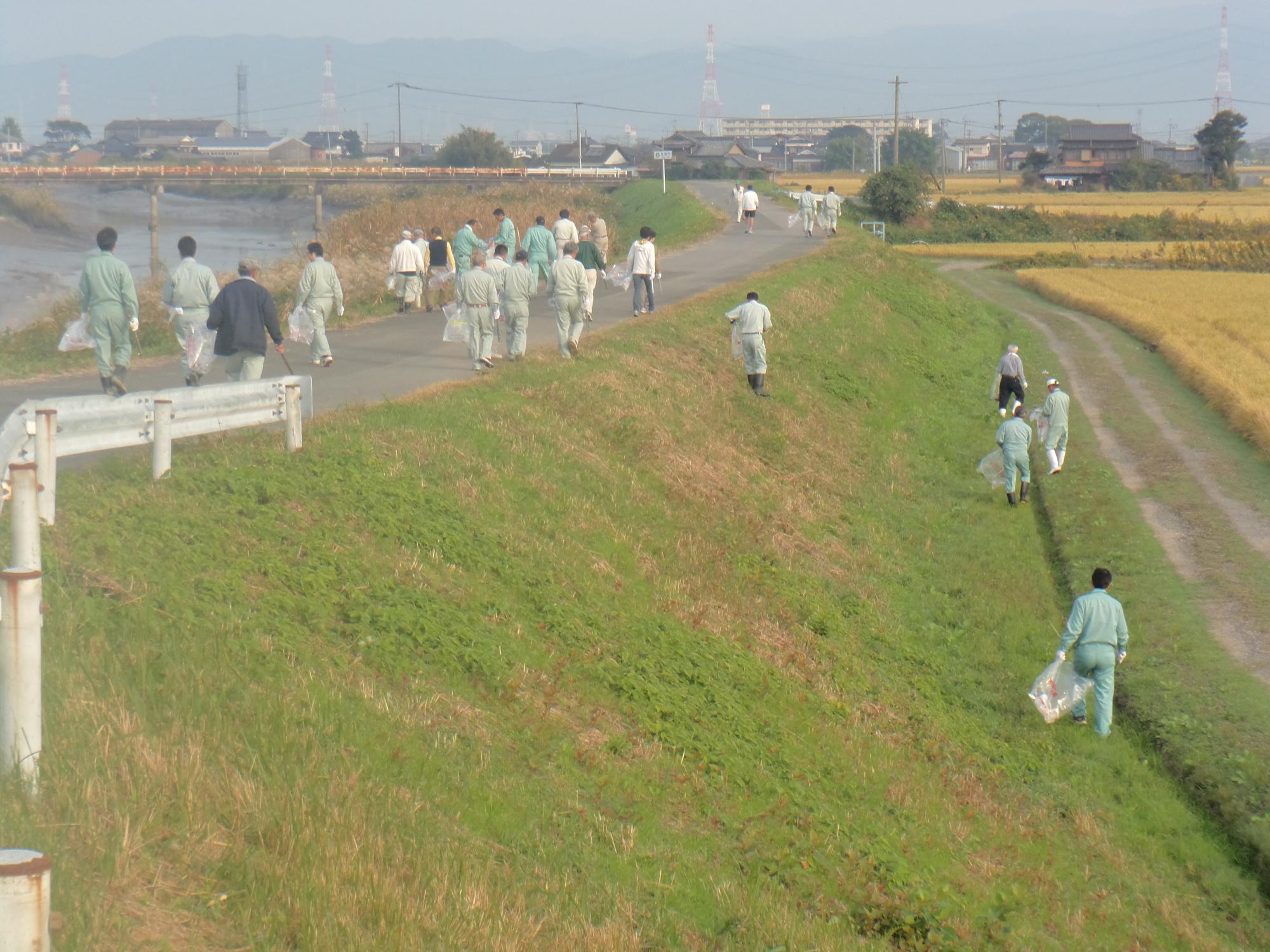 川沿いの広い土手で作業服姿の参加者たちが歩道や斜面に散らばってごみ拾いを協力して進める活動風景の写真