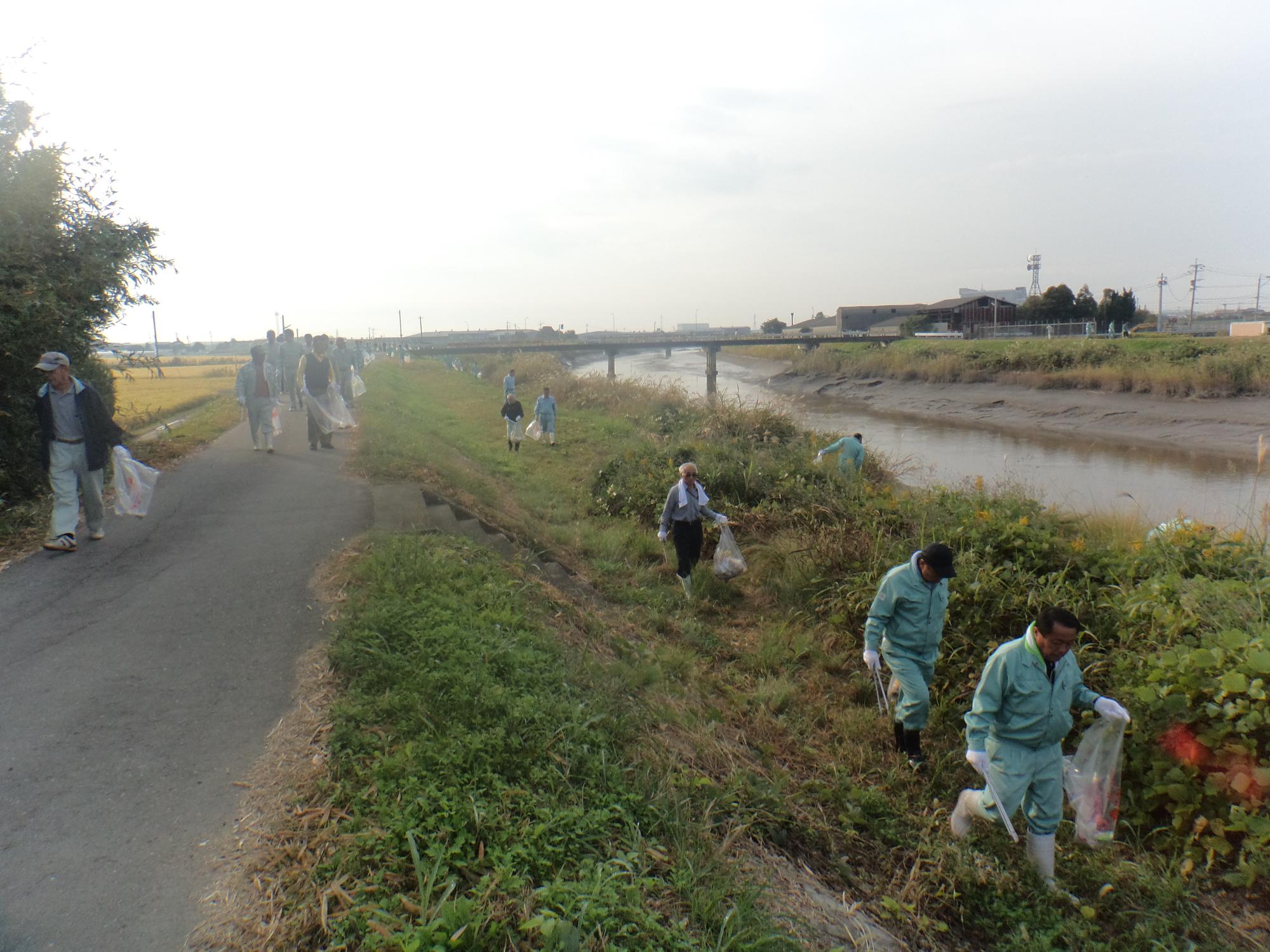 川沿いの土手で作業服姿の参加者たちがごみ袋を手に草むらや道沿いを清掃して回る様子の写真