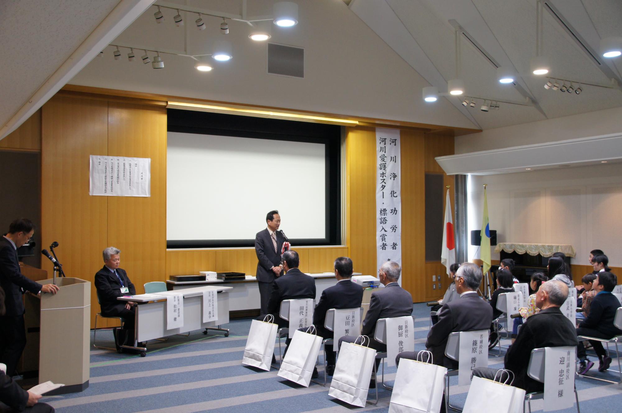 表彰式の会場で登壇者が前方で挨拶し、来賓が並んで着席し足元に贈呈品の袋が置かれた様子の写真