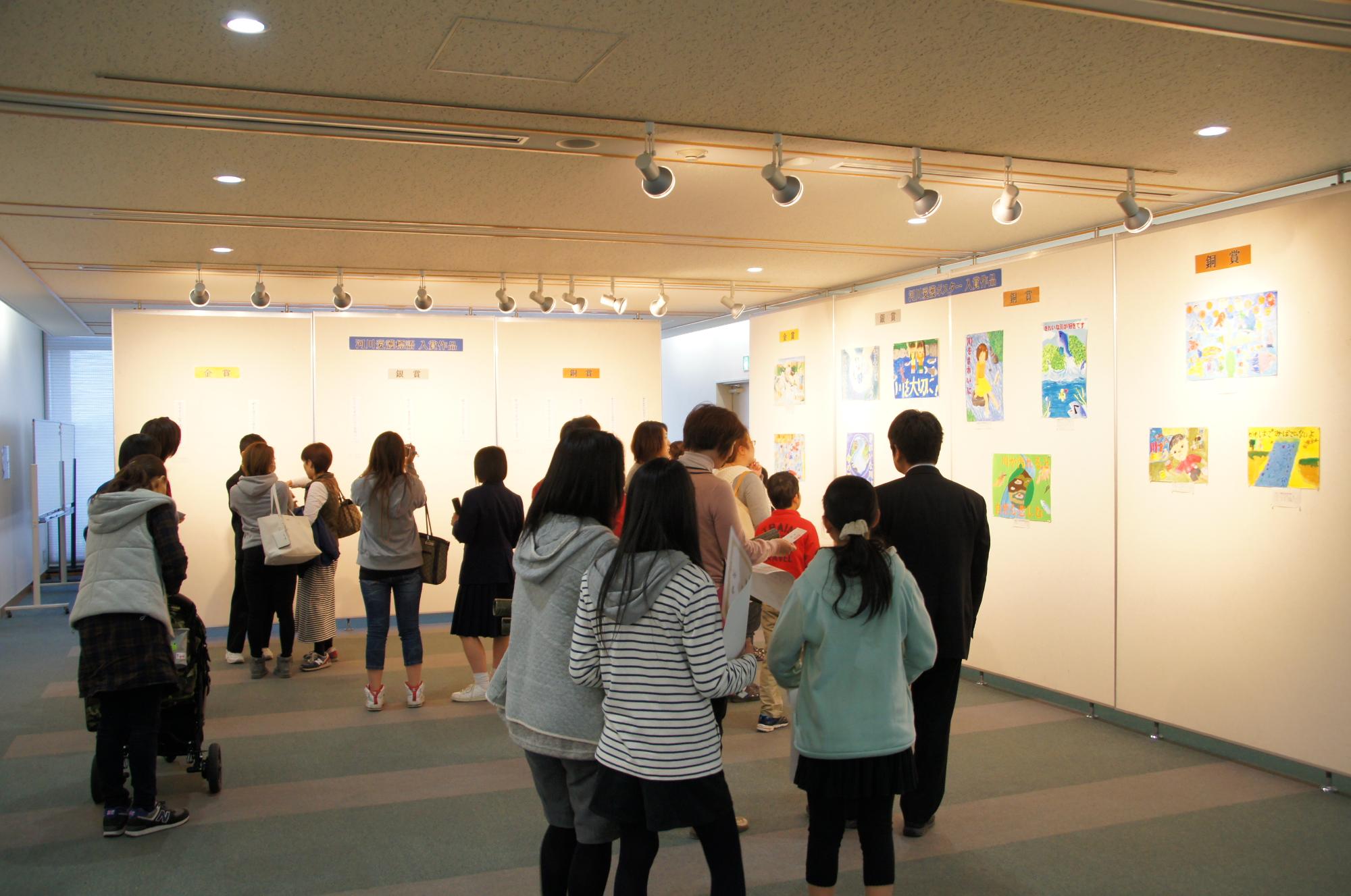 展示会場で来場者が壁に並ぶ色鮮やかな児童作品を眺めつつ歩き回る静かな鑑賞風景の写真