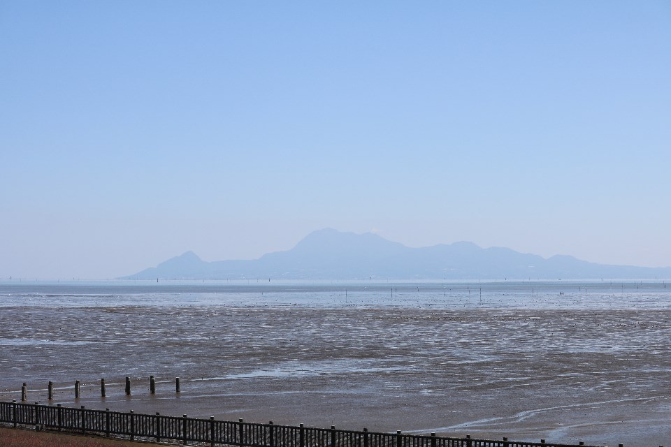 晴れ渡る青空の下、広大な干潟が手前に広がり、その奥に広がる海を越えて遠くに霞んだ山々が連なる、開放感あふれる穏やかな風景を捉えた写真
